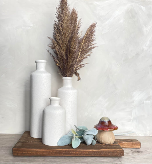 Decorative setup with white vases, dried pampas grass, and a small mushroom on a wooden board against a light background.