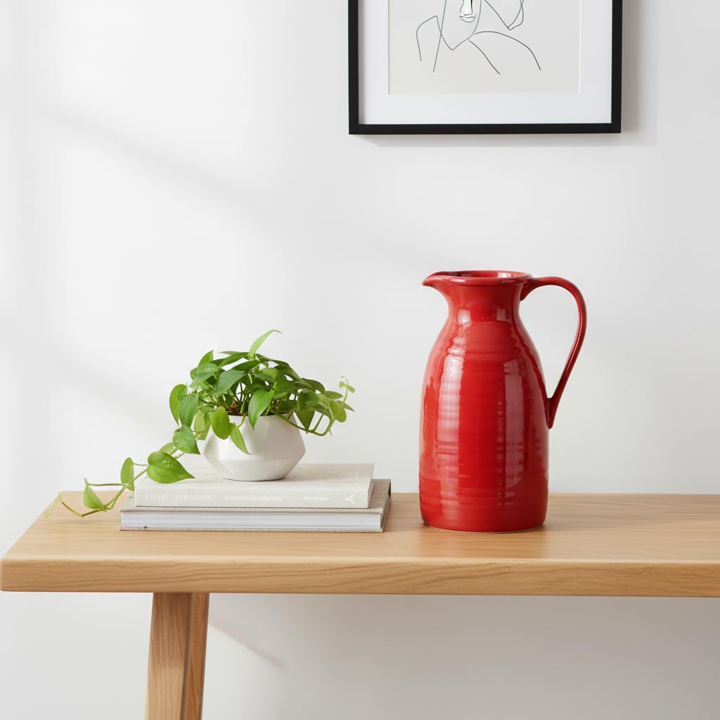 Red pitcher on a wooden table with a plant and books, against a white wall with a framed picture.