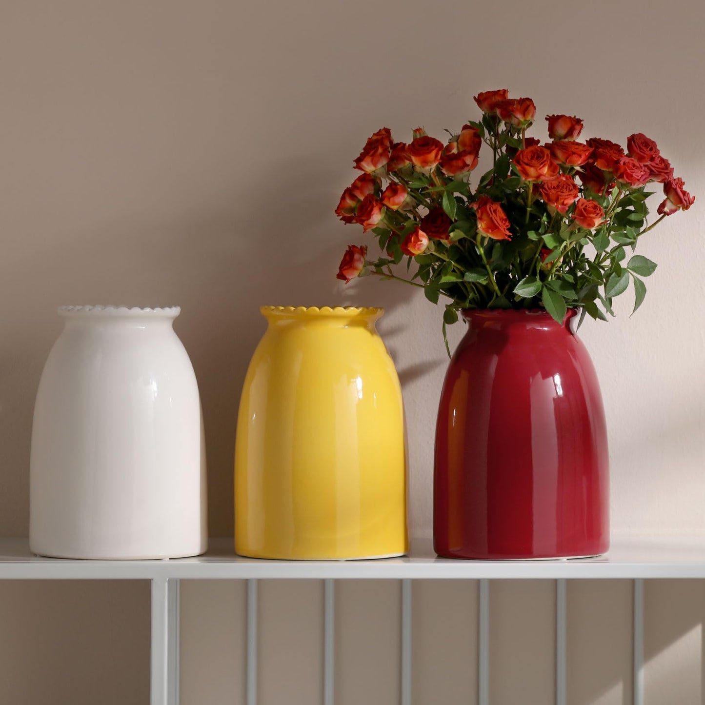 Three vases of different colors (white, yellow, red) with a red flower arrangement on a shelf.