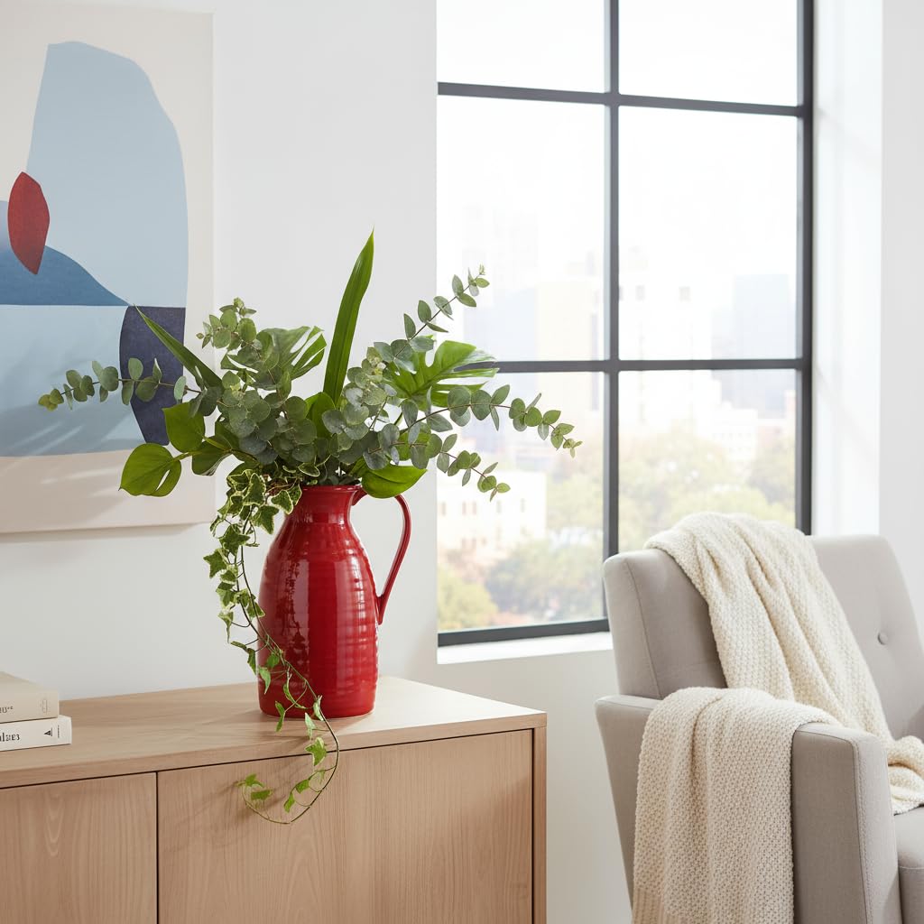 Red vase with greenery on a wooden cabinet in a living room with a window and sofa.