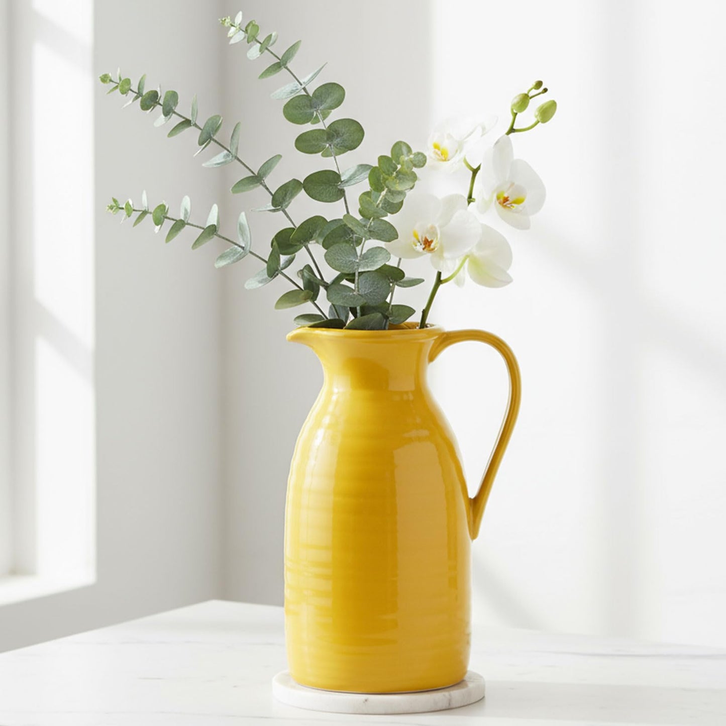 Yellow pitcher with greenery and white flowers on a light surface with a neutral background