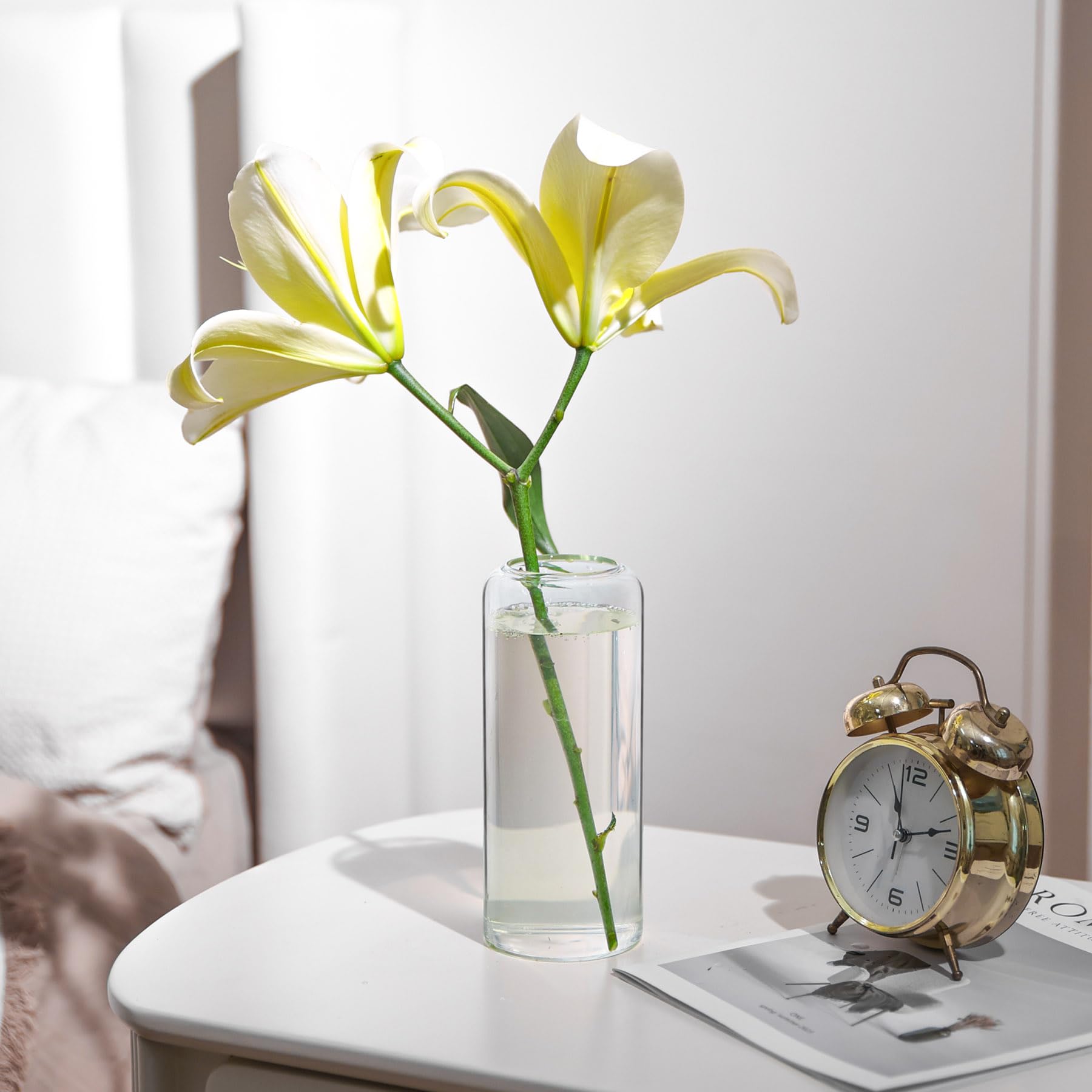 Yellow lily in a clear vase on a table with a gold alarm clock and white surface.
