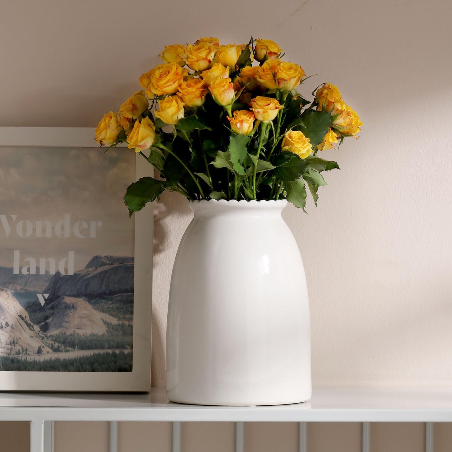 White vase with yellow roses on a shelf next to a framed picture