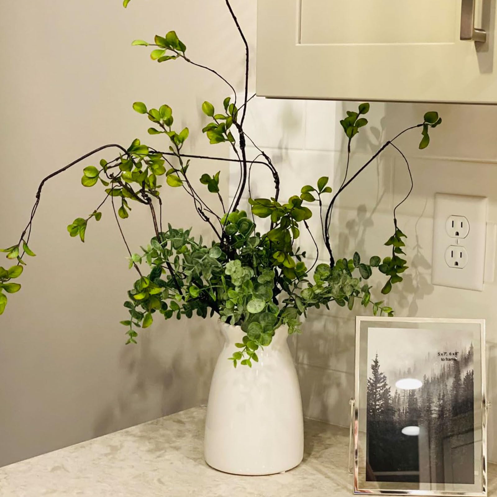 White vase with greenery on a table next to a framed picture