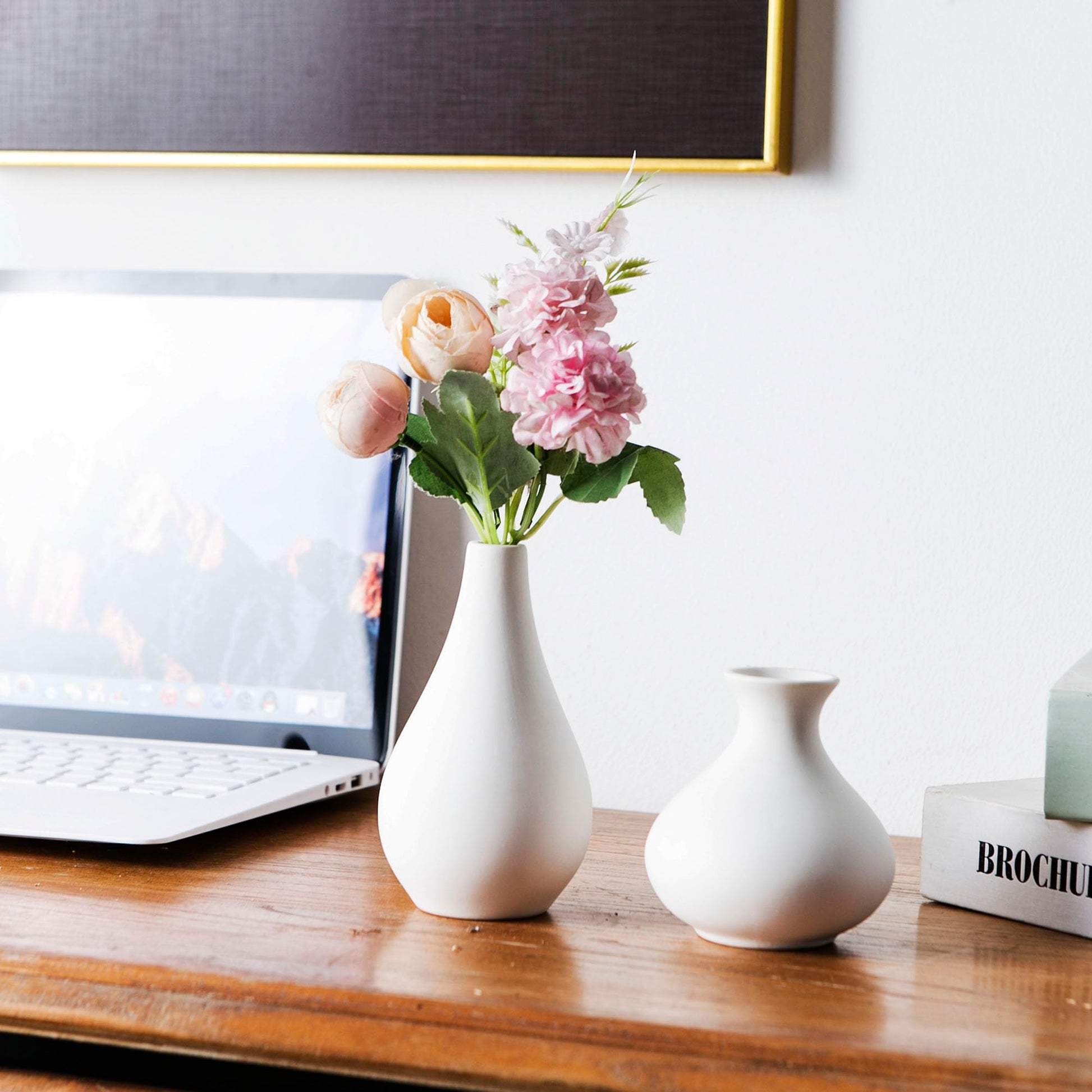 White vases with pink flowers on a wooden desk next to a laptop