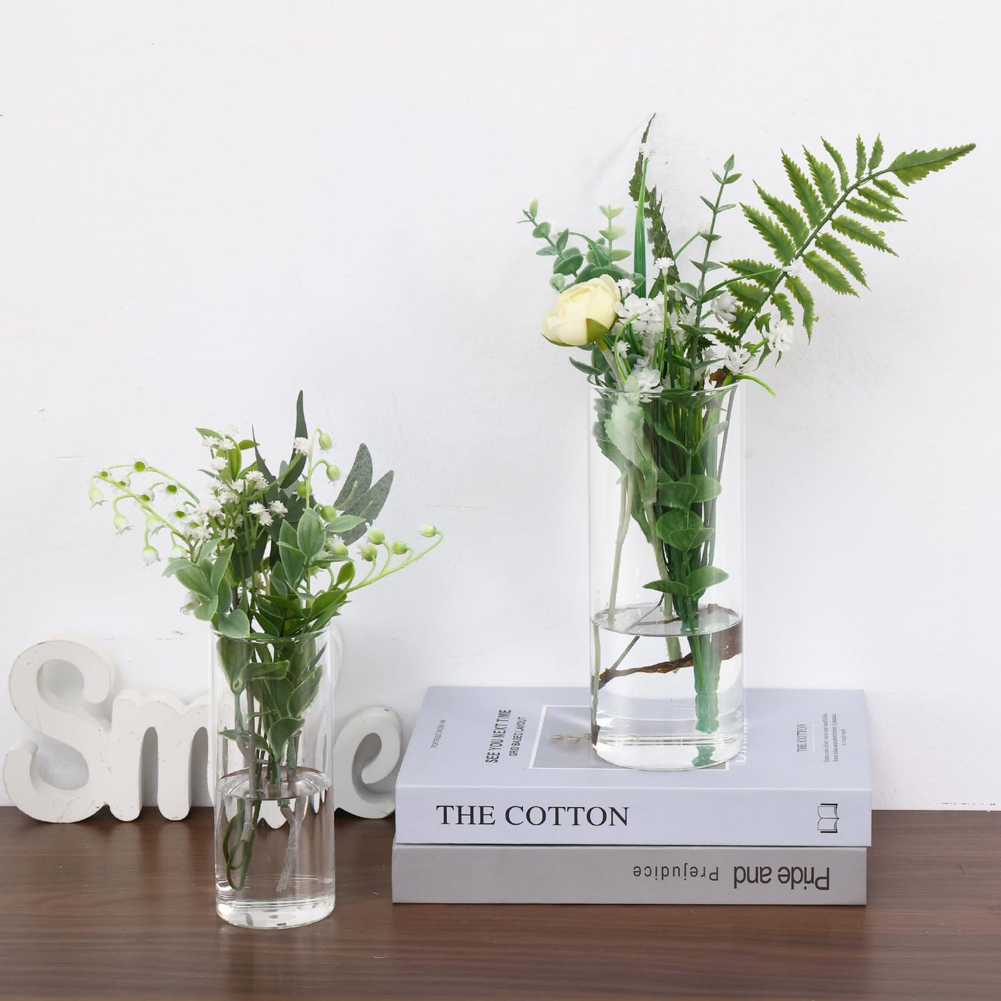 Two glass vases with greenery on a wooden surface with a white background
