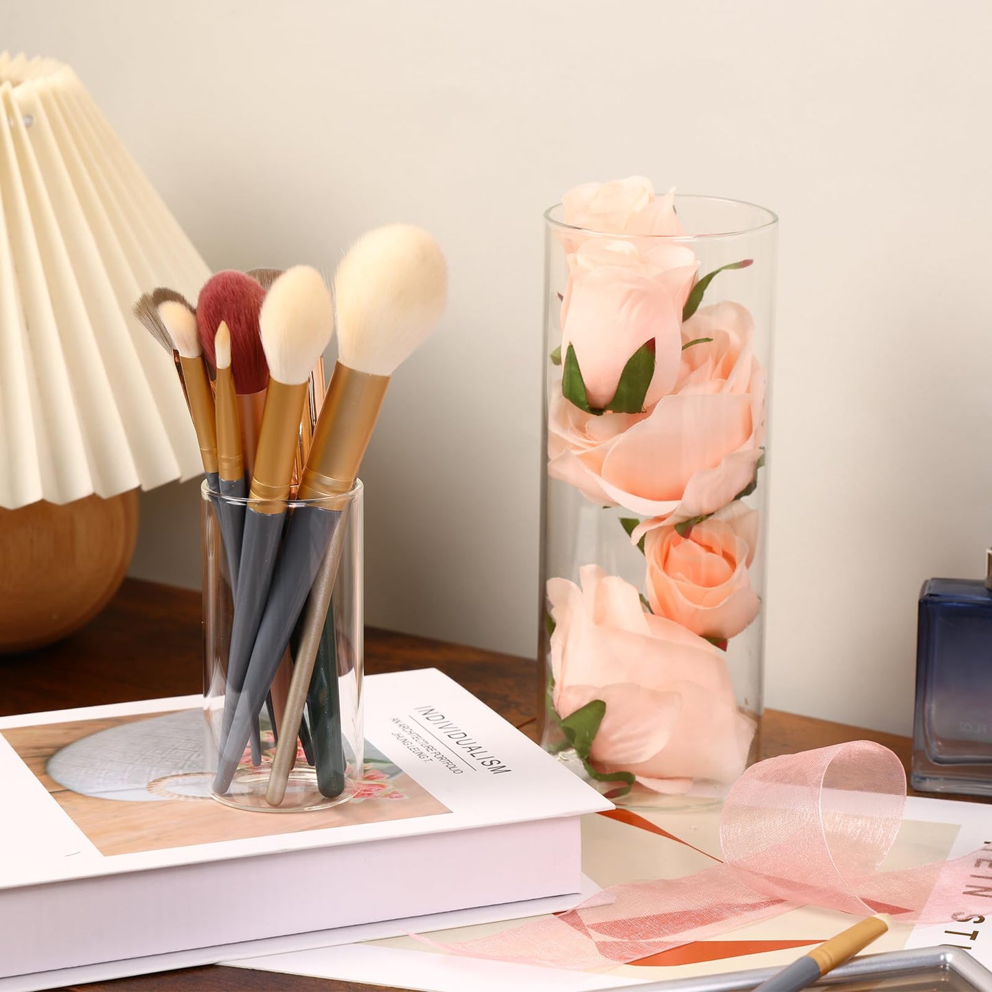 Makeup brushes in a glass holder next to a vase of pink roses on a table.