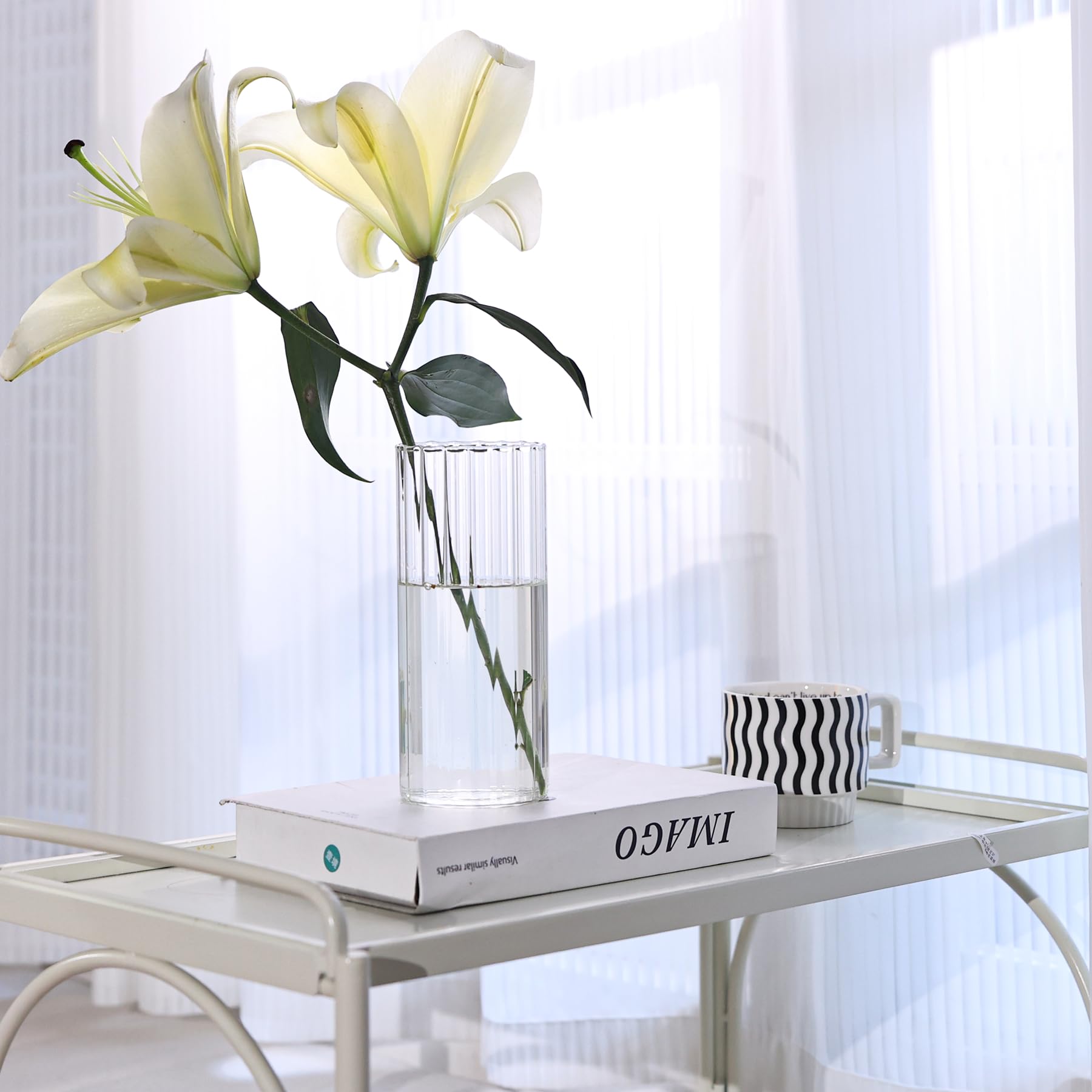 Clear glass vase with white lilies on a white table with a book and black and white patterned mug.