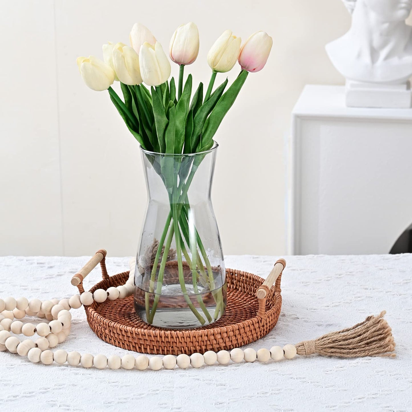 Clear vase with white and pink tulips on a woven tray with decorative beads and a broomstick on a white surface.