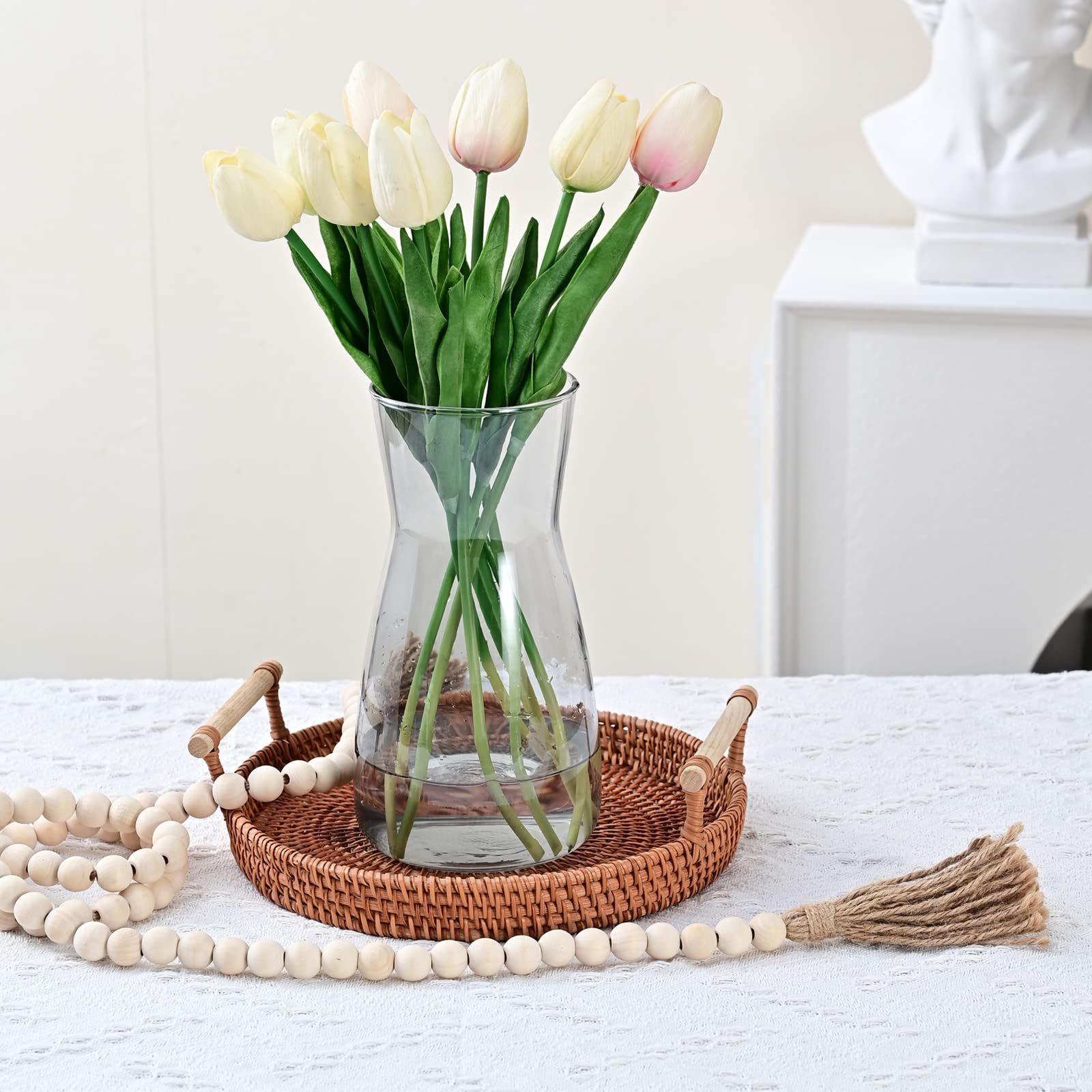 Clear vase with white and pink tulips on a woven tray with decorative beads and a broomstick on a white surface.