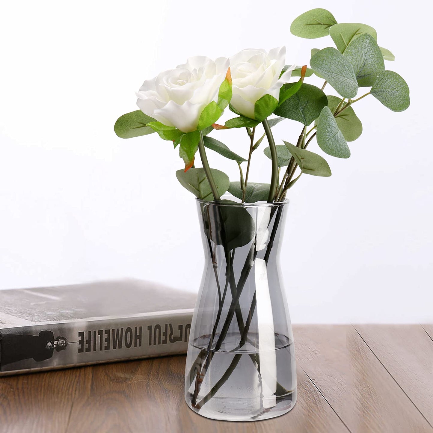 Clear glass vase with white flowers and green leaves on a wooden surface.