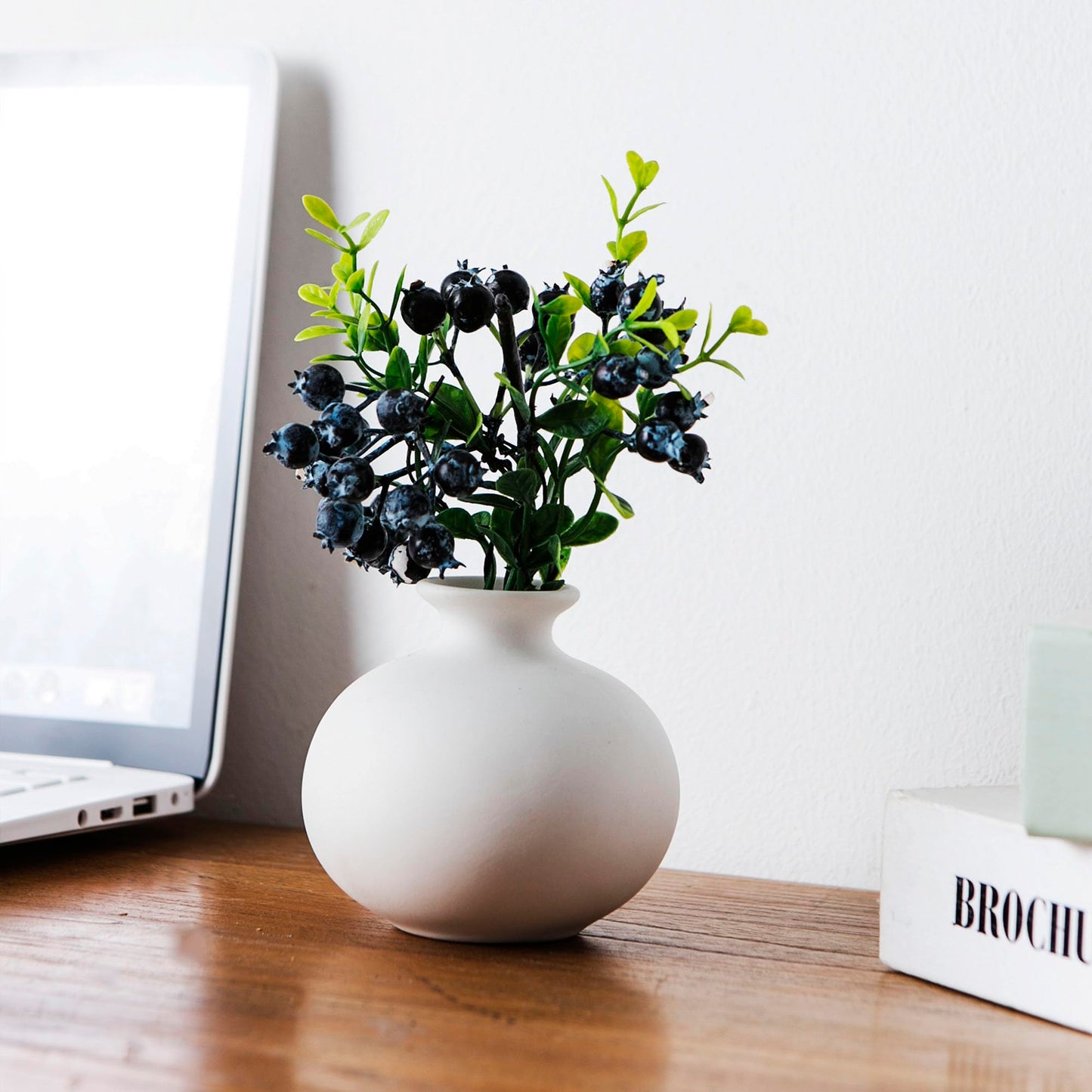 White vase with greenery on a wooden surface next to a laptop