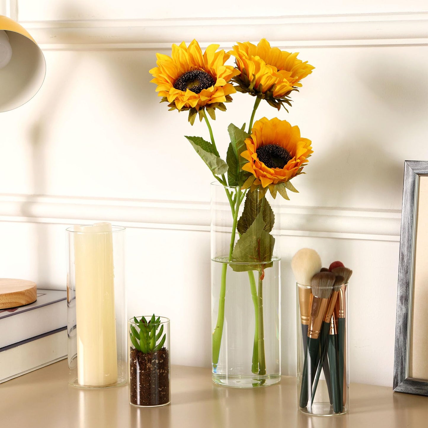 Sunflowers in a clear vase on a table with candles and makeup brushes.