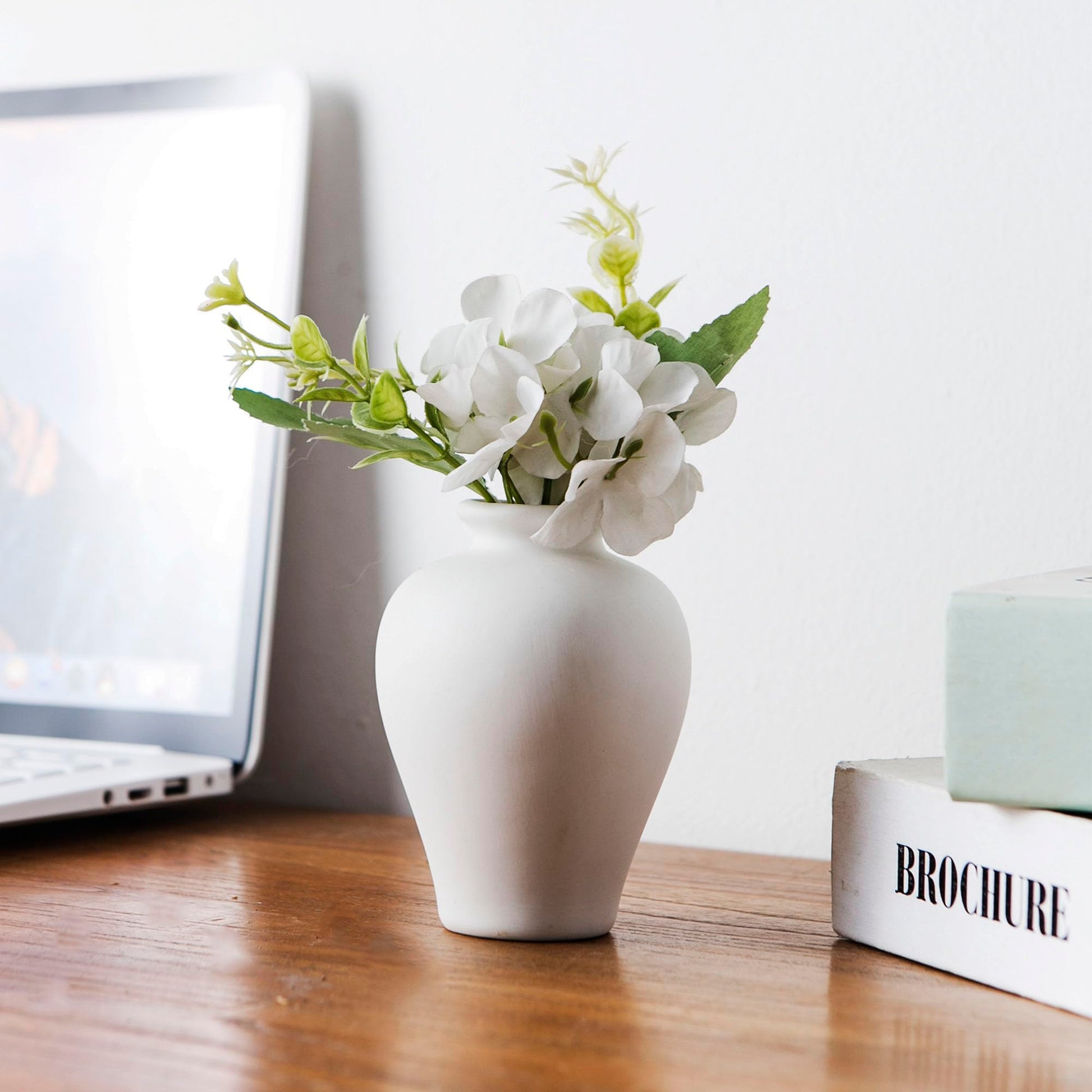 White vase with flowers on a wooden desk next to a laptop and books