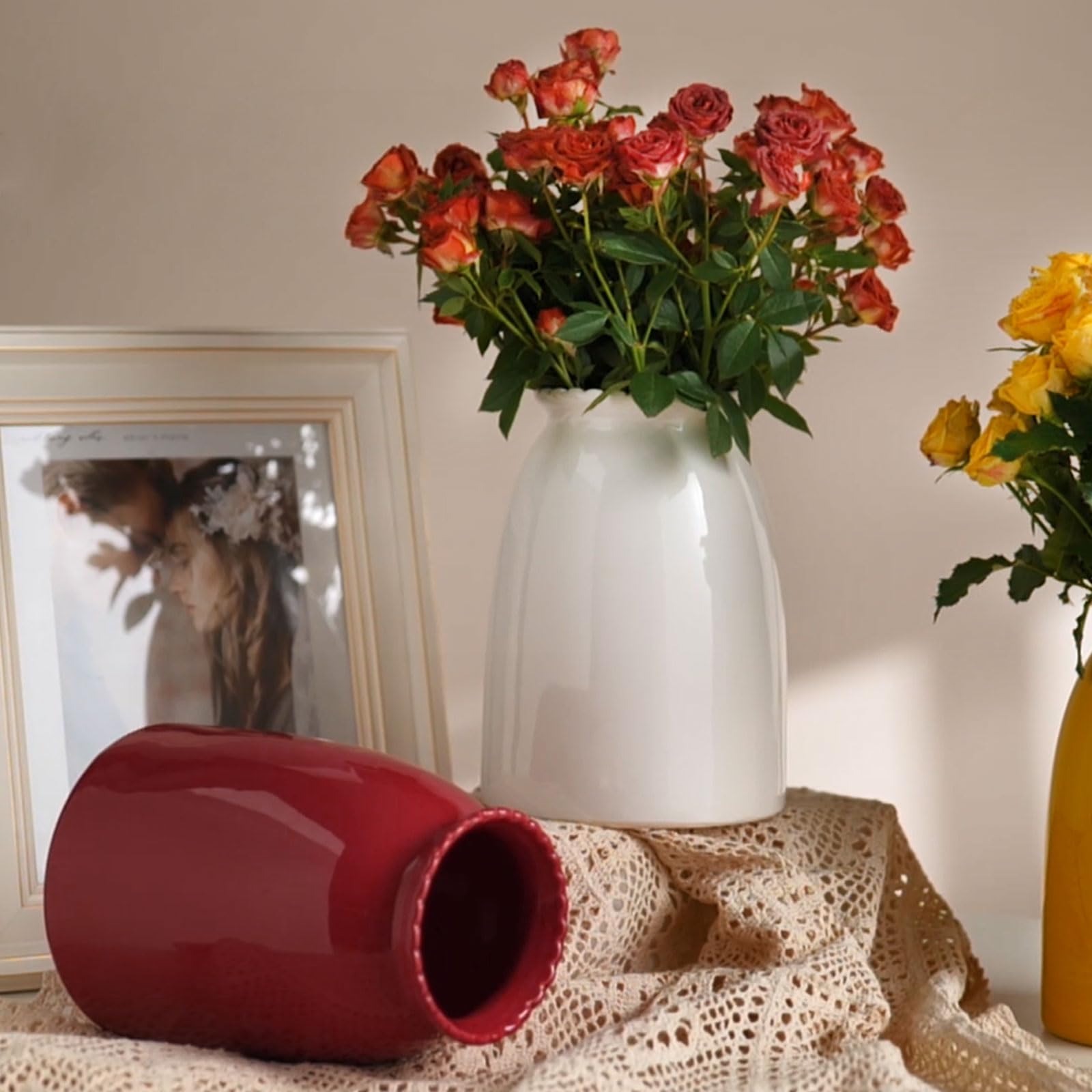 White vase with red and orange flowers on a textured surface with a blurred photo frame in the background.