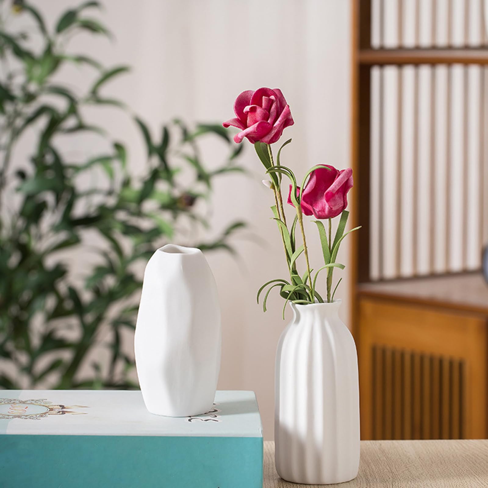 White vase with pink flowers on a table next to a green box, with a blurred plant and wooden shelf in the background.