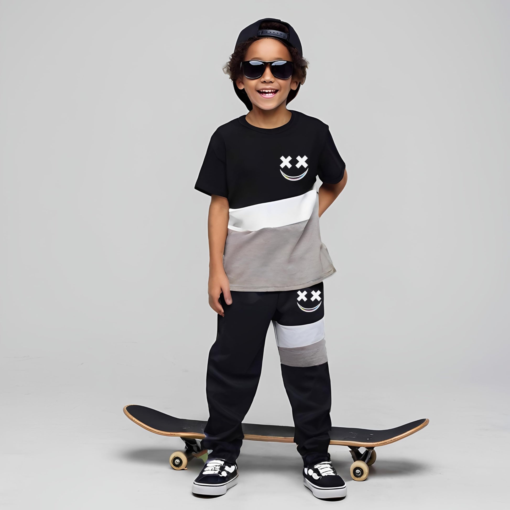 Child wearing a black and gray outfit with smiley face designs, standing with a skateboard on a plain background.
