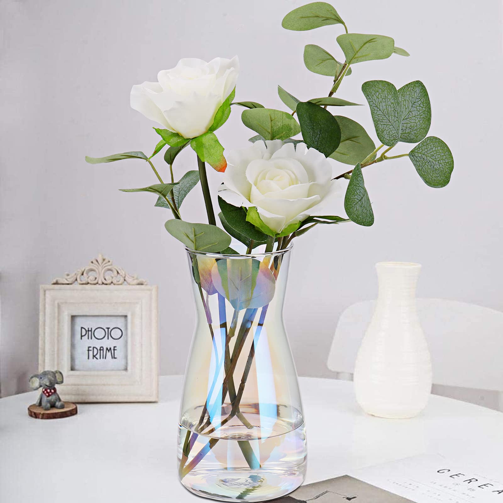 Clear glass vase with white flowers and green leaves on a white surface.