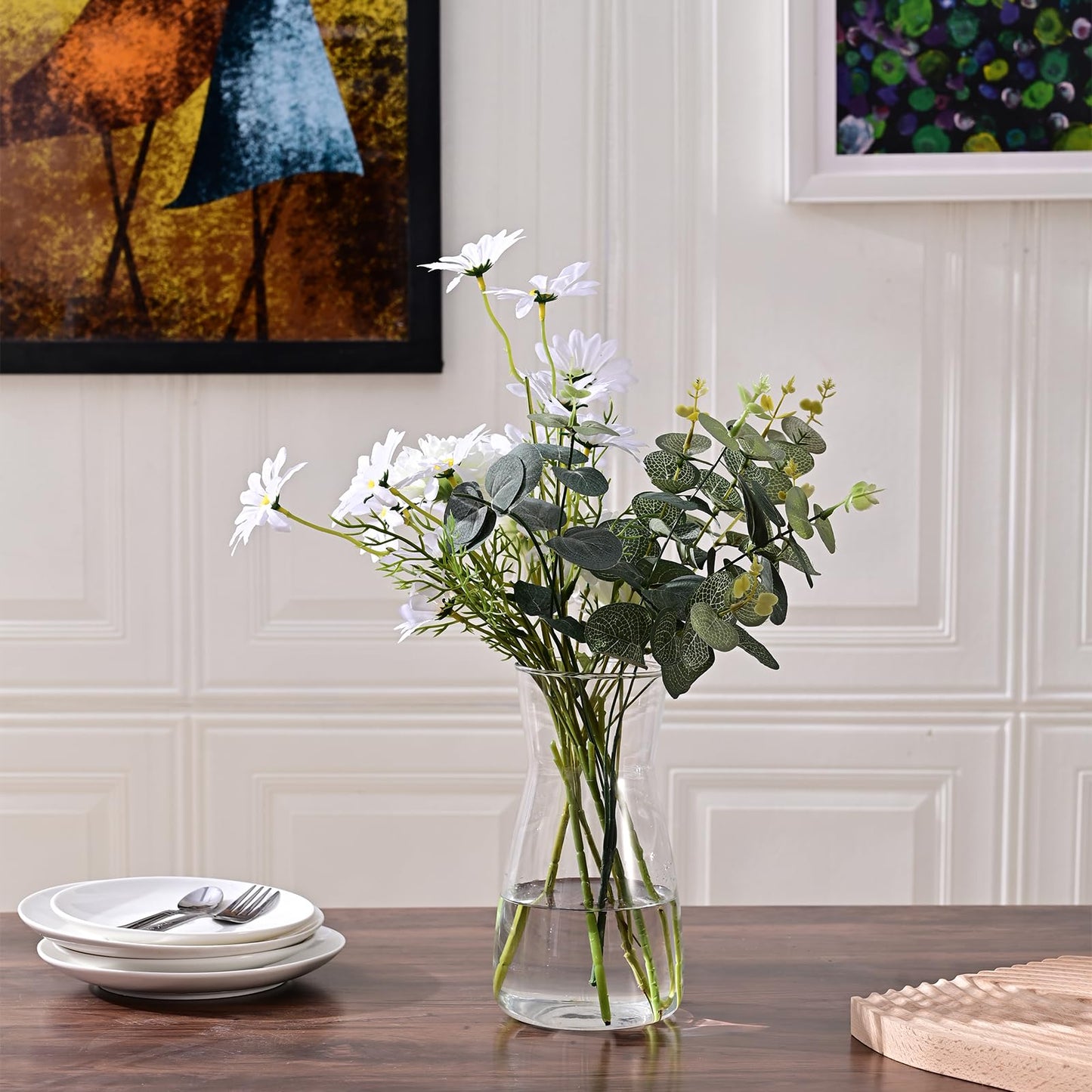 Clear glass vase with white flowers and green leaves on a wooden table against a wall with framed artwork.