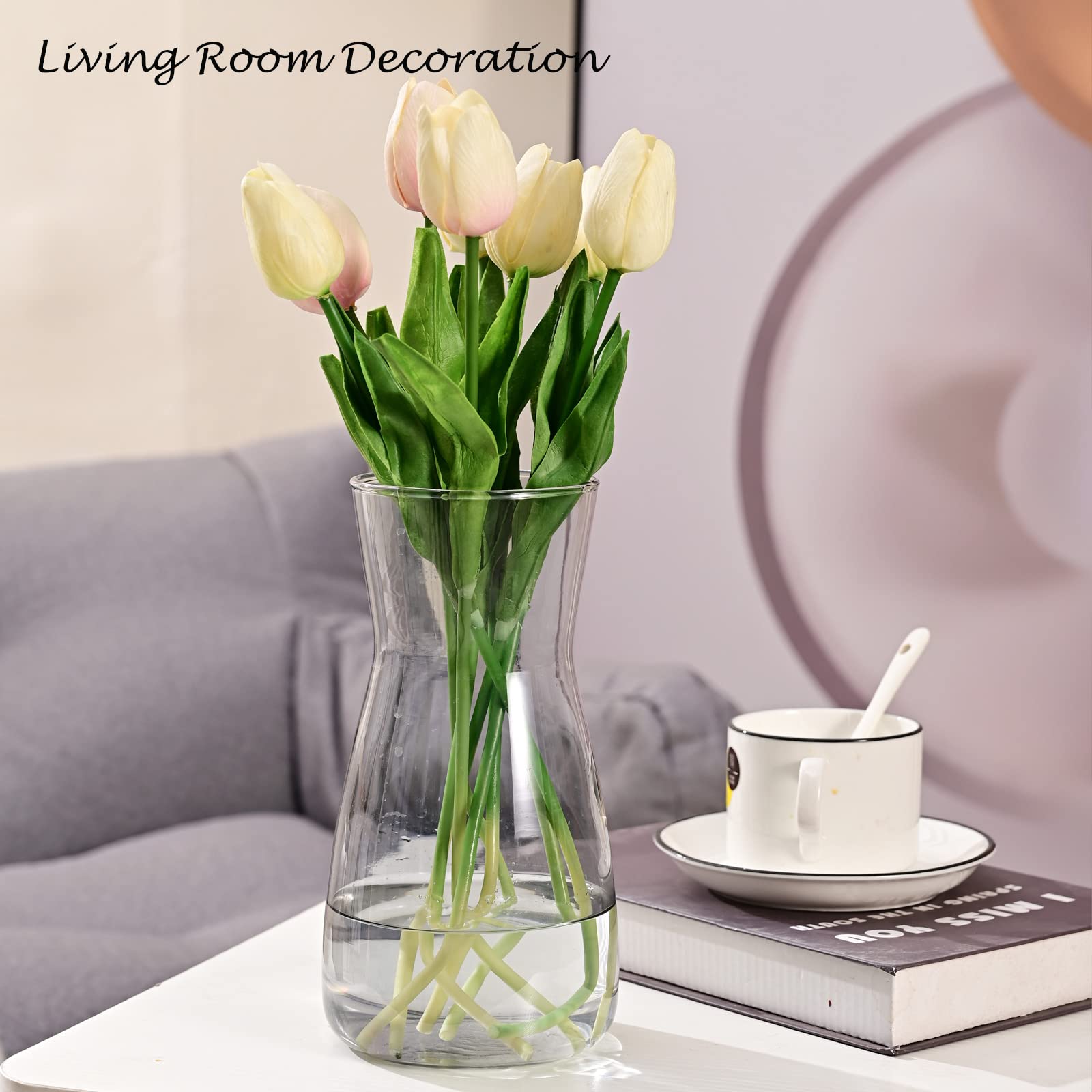 Clear vase with pink tulips on a table next to a book and cup in a living room setting.