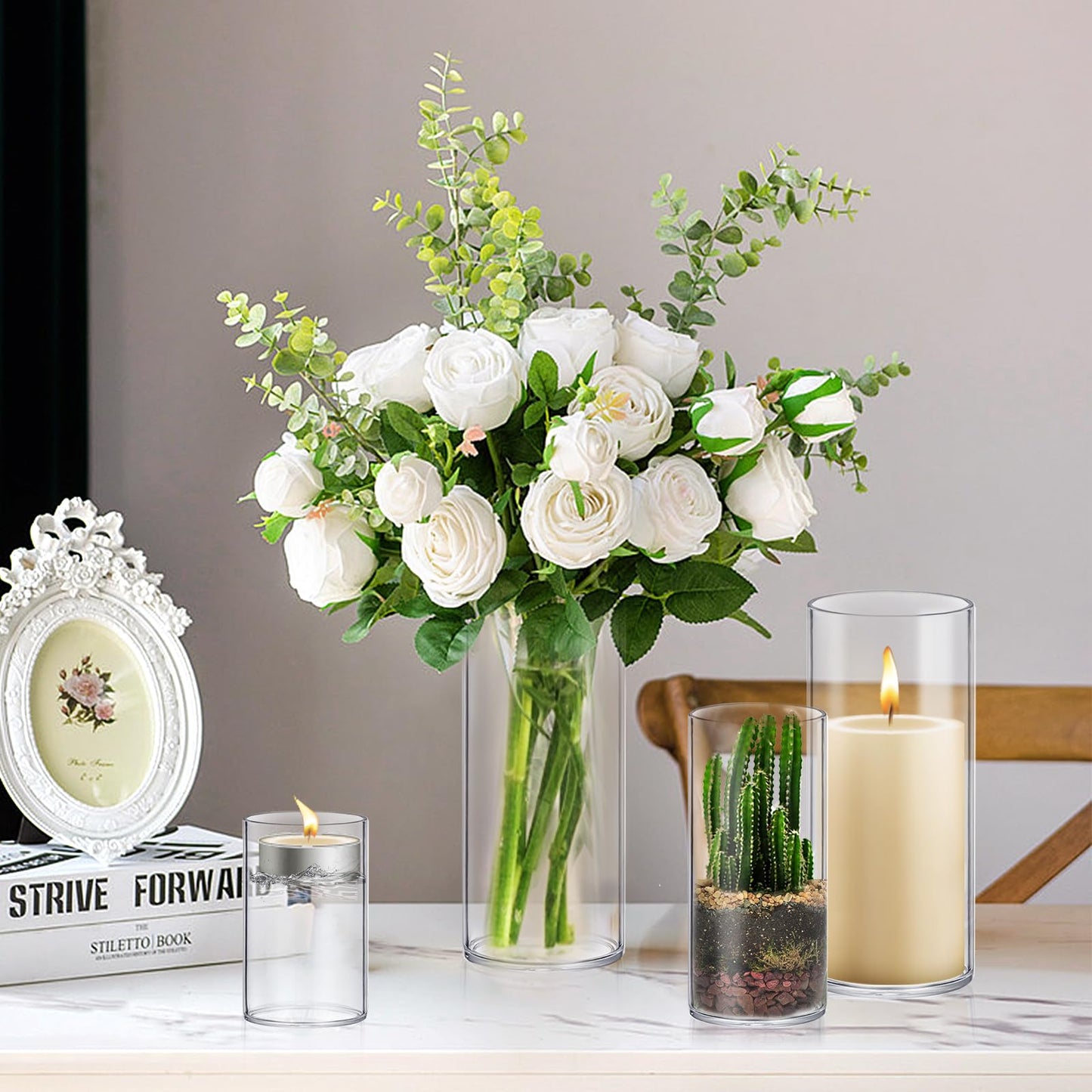 Bouquet of white roses and greenery in a vase with candles and a framed picture on a table.