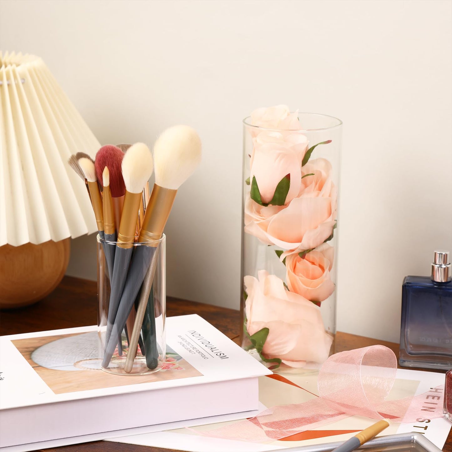 Makeup brushes in a glass holder on a table with a vase of flowers and a book.