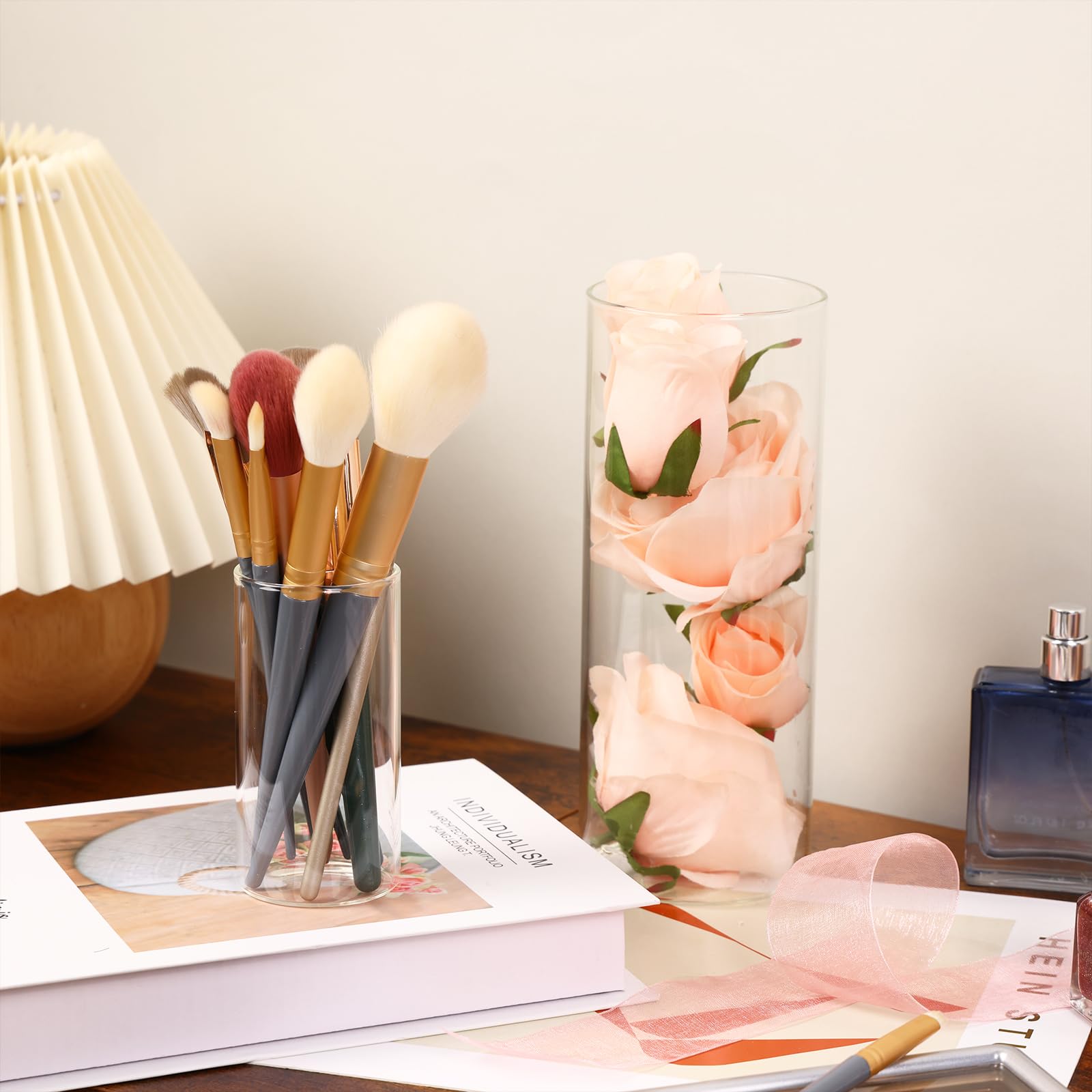 Makeup brushes in a glass holder on a table with a vase of flowers and a book.