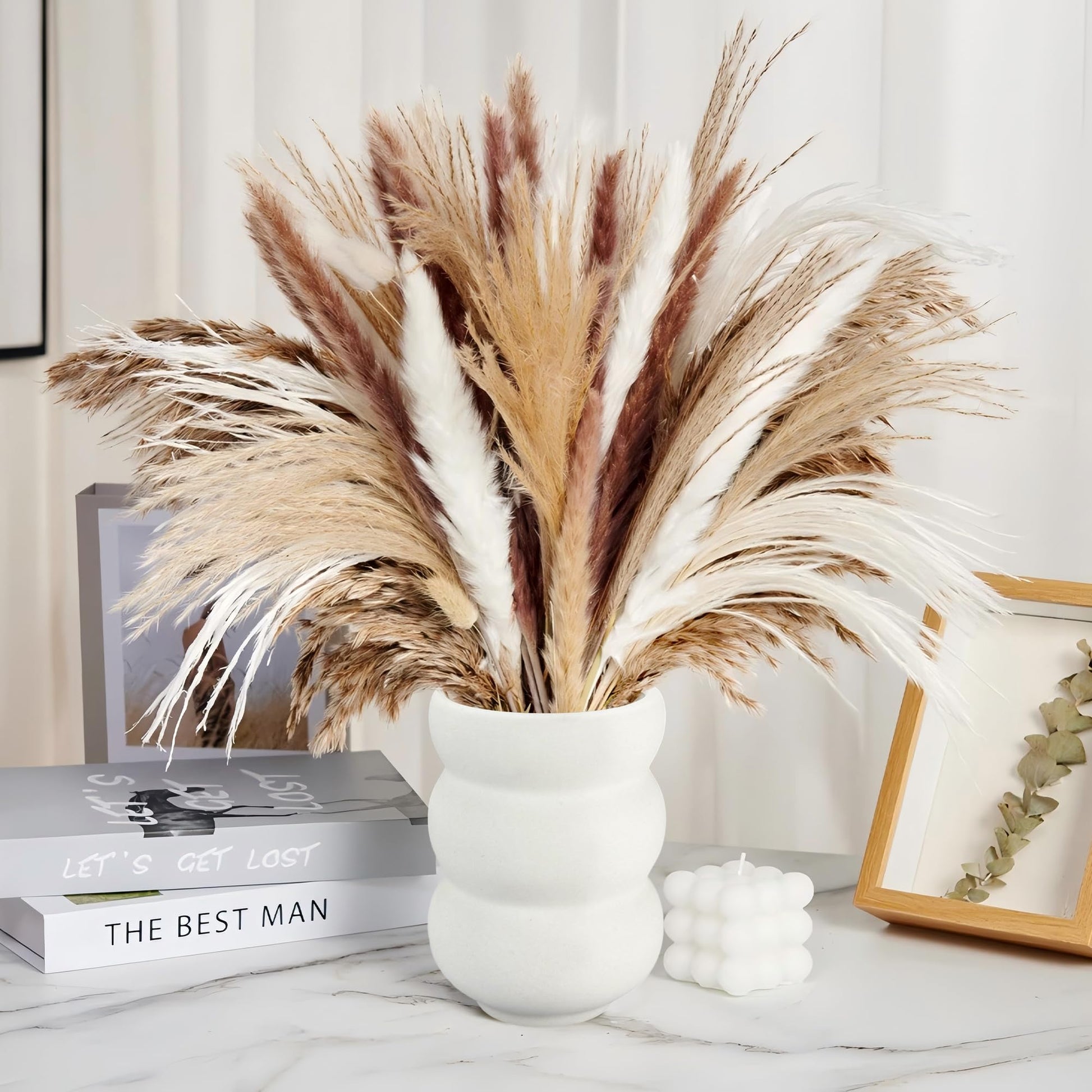 Decorative arrangement of pampas grass in a white vase on a table with books and a candle.