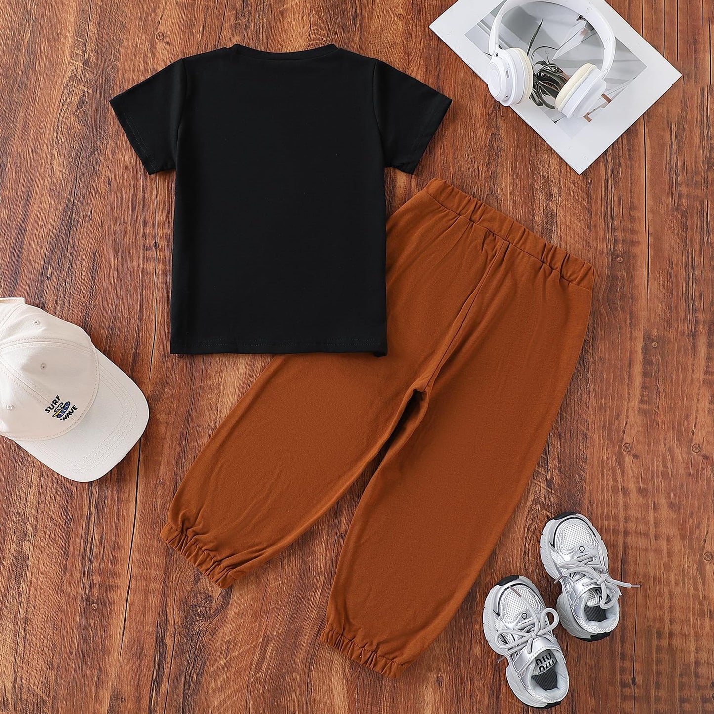 Black t-shirt, brown pants, white sneakers, and cap on a wooden surface with a magazine and headphones.