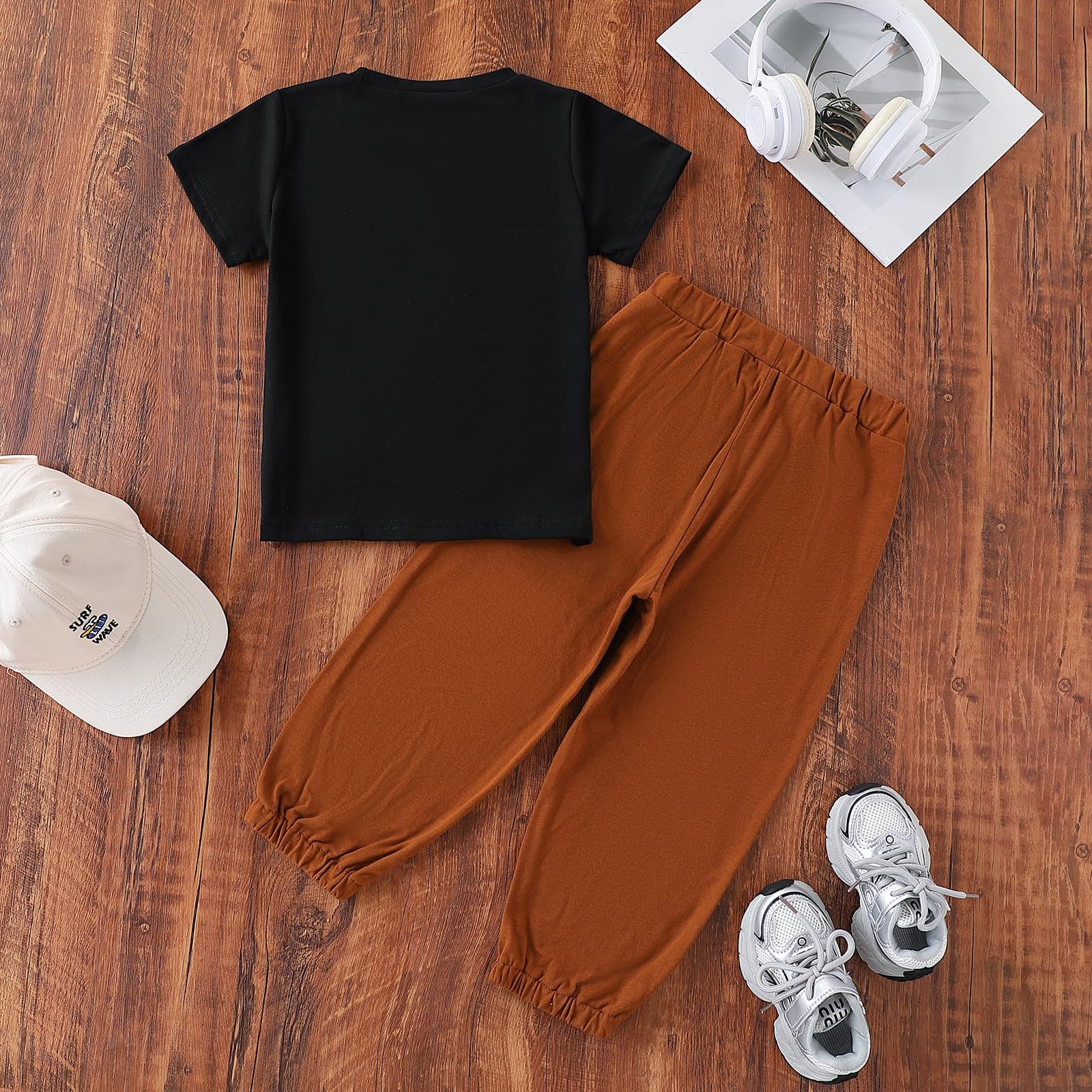 Black t-shirt, brown pants, white sneakers, and cap on a wooden surface with a magazine and headphones.