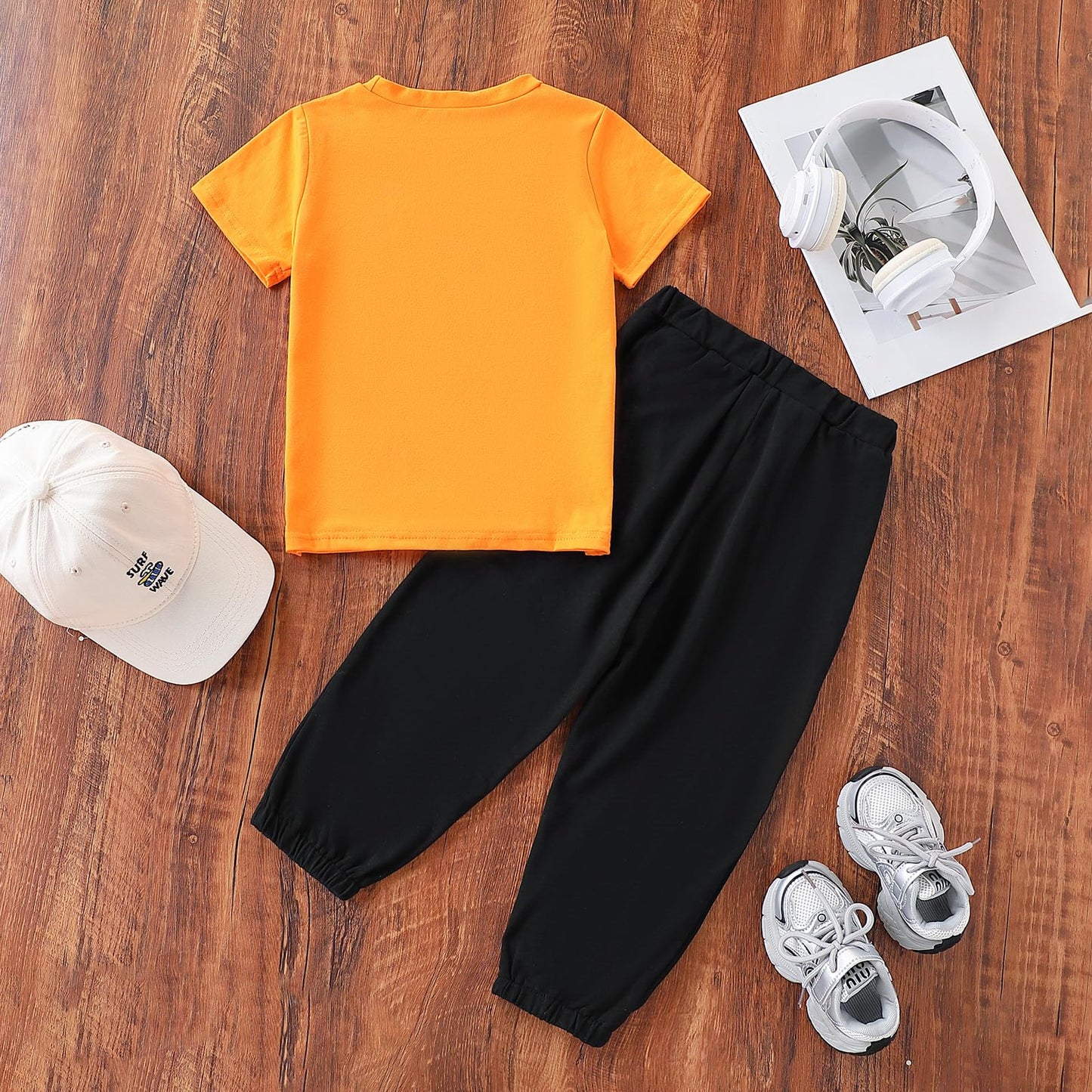 Orange t-shirt, black pants, white sneakers, and cap on a wooden surface with a photo album.