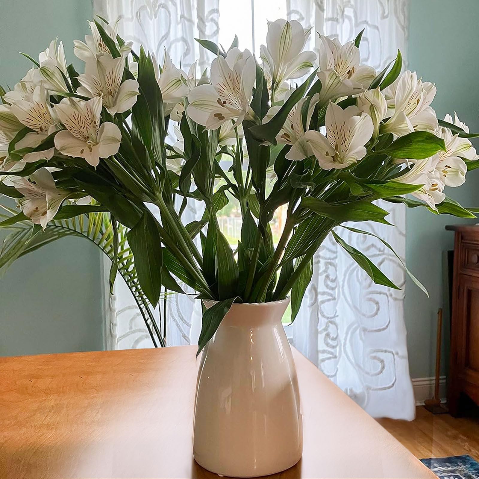 White flowers in a vase on a wooden table with a light blue wall and white lace curtain in the background.