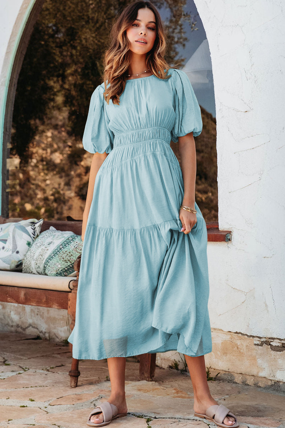 Woman wearing a light blue dress standing in front of a white wall and wooden bench.